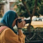 Female photographer with camera capturing street scenes, wearing casual attire and headscarf.