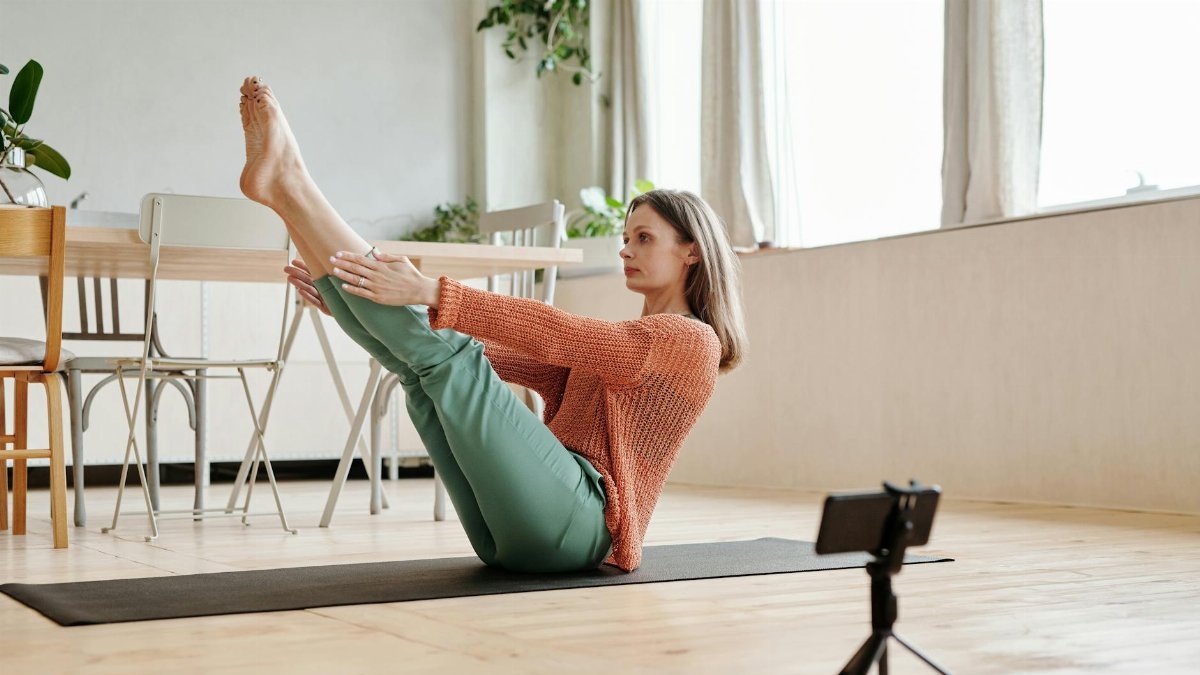Woman doing a yoga pose indoors, using a smartphone to record or follow along.