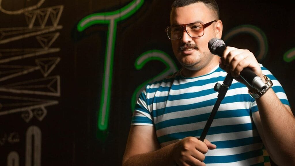 Man holding a microphone doing stand-up comedy in casual striped shirt on stage.