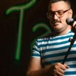 Man holding a microphone doing stand-up comedy in casual striped shirt on stage.