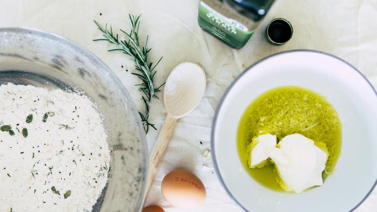 Flat lay of baking ingredients including flour, eggs, and olive oil with rosemary on wooden table.