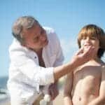 A father applies sunscreen to his smiling son on a sunny beach day.