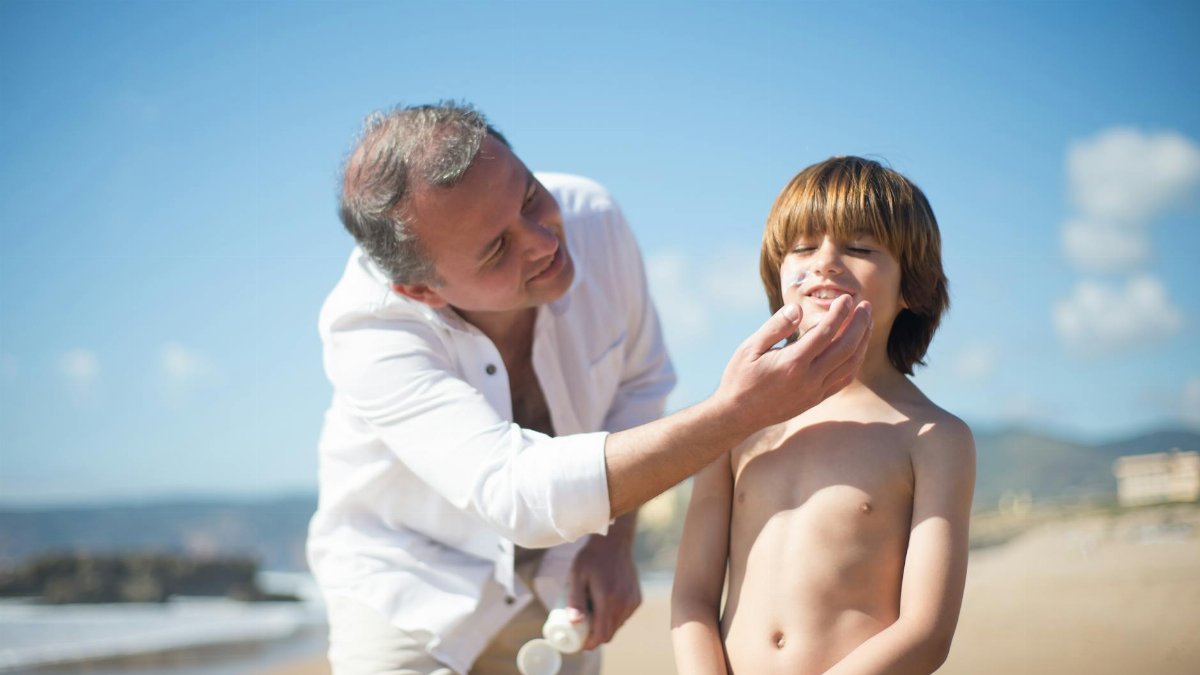 A father applies sunscreen to his smiling son on a sunny beach day.
