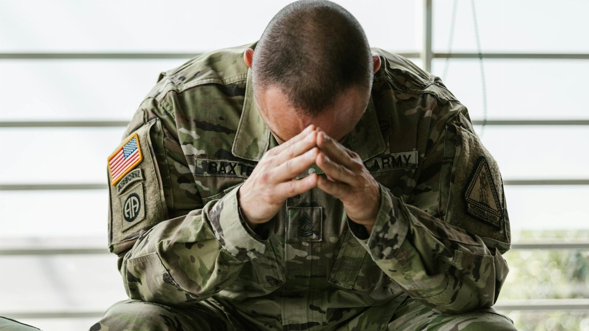 A soldier in camouflage sitting indoors, appearing stressed and overwhelmed.