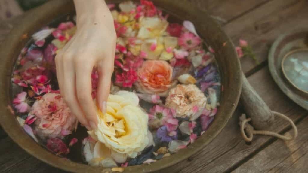 A hand gently arranges colorful flower petals floating in a rustic wooden bowl.