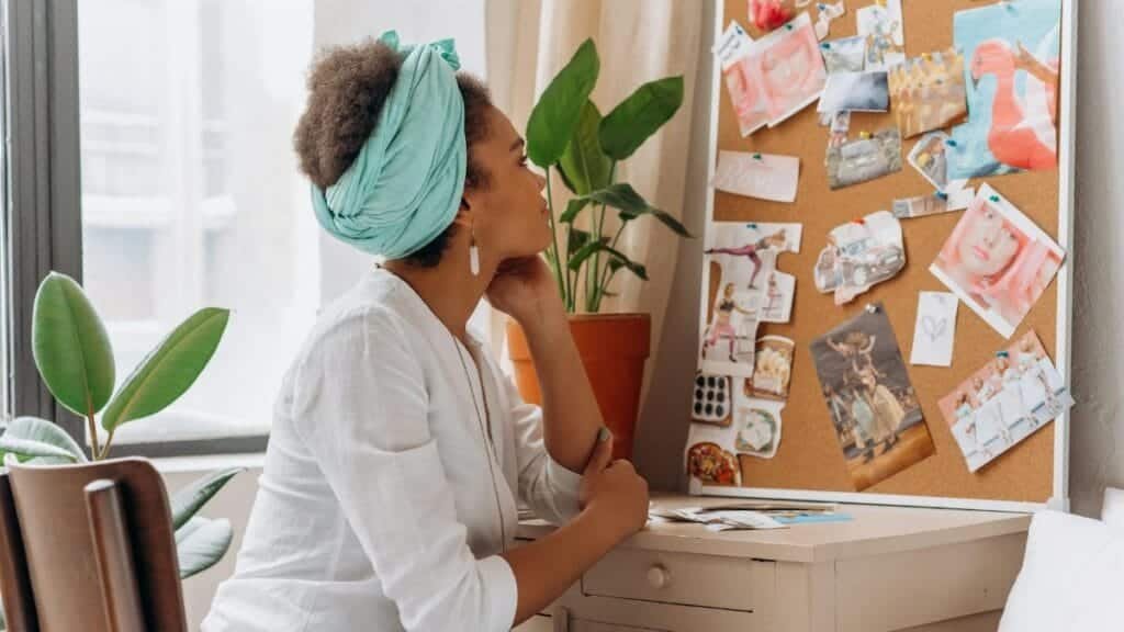 African American woman reflecting on a vision board by a window with plants.