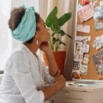 African American woman reflecting on a vision board by a window with plants.