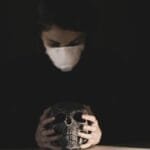 A person wearing a mask holds a skull under a spotlight in a dark studio, symbolizing mortality and protection.