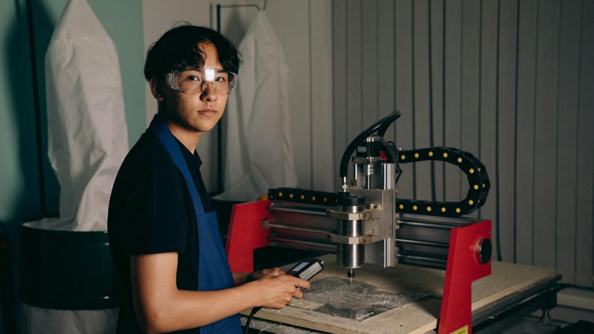 Portrait of a young worker operating a CNC machine in a workshop, wearing safety glasses and an apron.