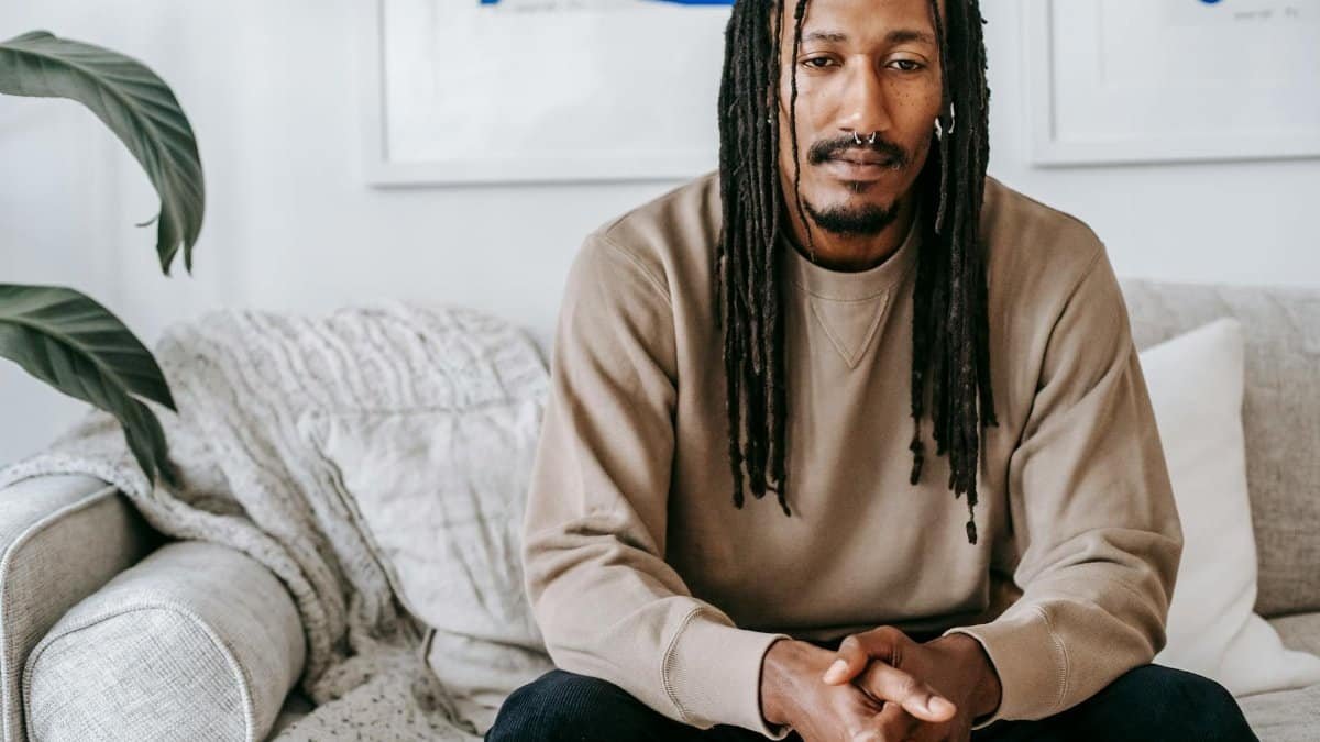 Crop thoughtful African American male in casual sweater with dreadlocks sitting with hands clasped on comfy couch in modern living room
