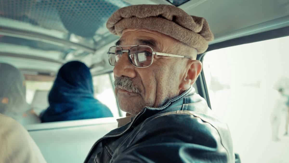 Elderly man with glasses and hat in a vehicle interior, looking back at the camera.
