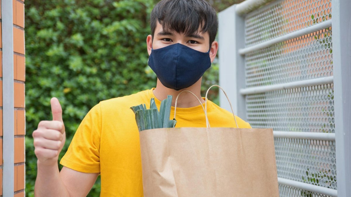 Young man in a mask holds groceries and gives a thumbs up at a metal gate outdoors.