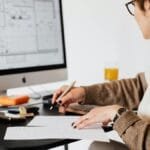 Woman designing architectural blueprints using desktop computer in a modern office setting.