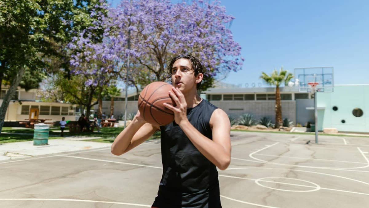A man in activewear practicing basketball on an outdoor court with trees and clear sky.