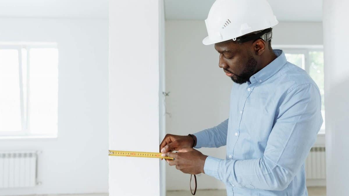 Construction worker measuring a wall indoors, wearing a white hard hat and blue shirt.