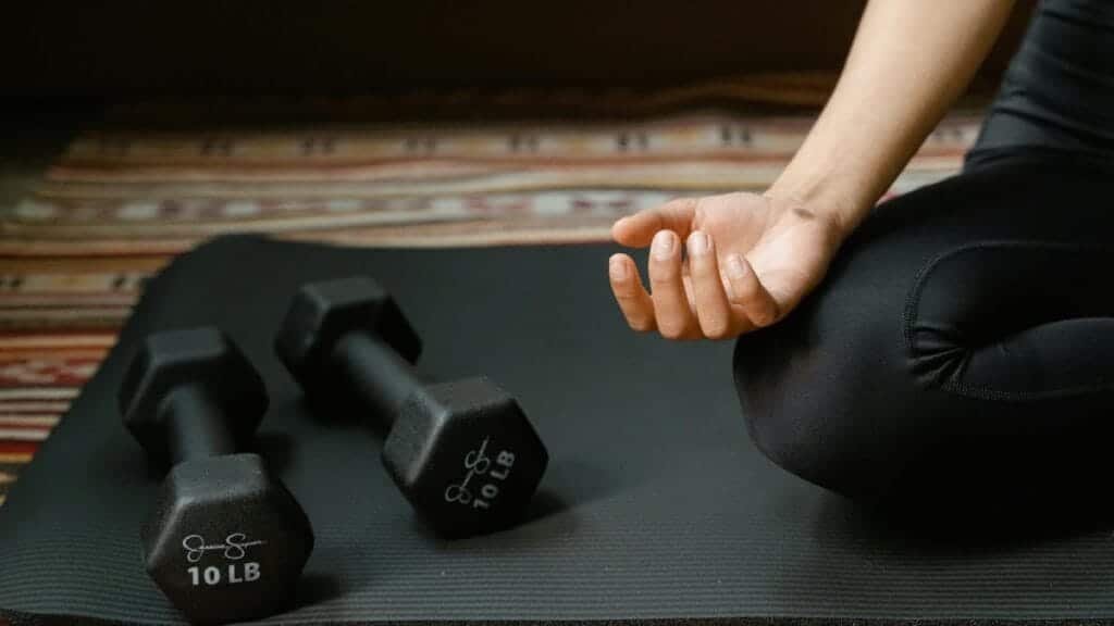 Close-up of a person's hand meditating next to dumbbells on a yoga mat, symbolizing fitness and mindfulness.