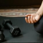 Close-up of a person's hand meditating next to dumbbells on a yoga mat, symbolizing fitness and mindfulness.