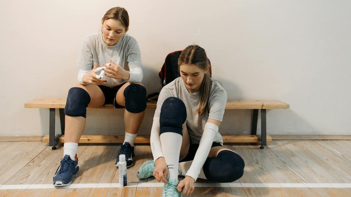 Two female volleyball players in sports gear prepare for a game, sitting on a wooden bench.