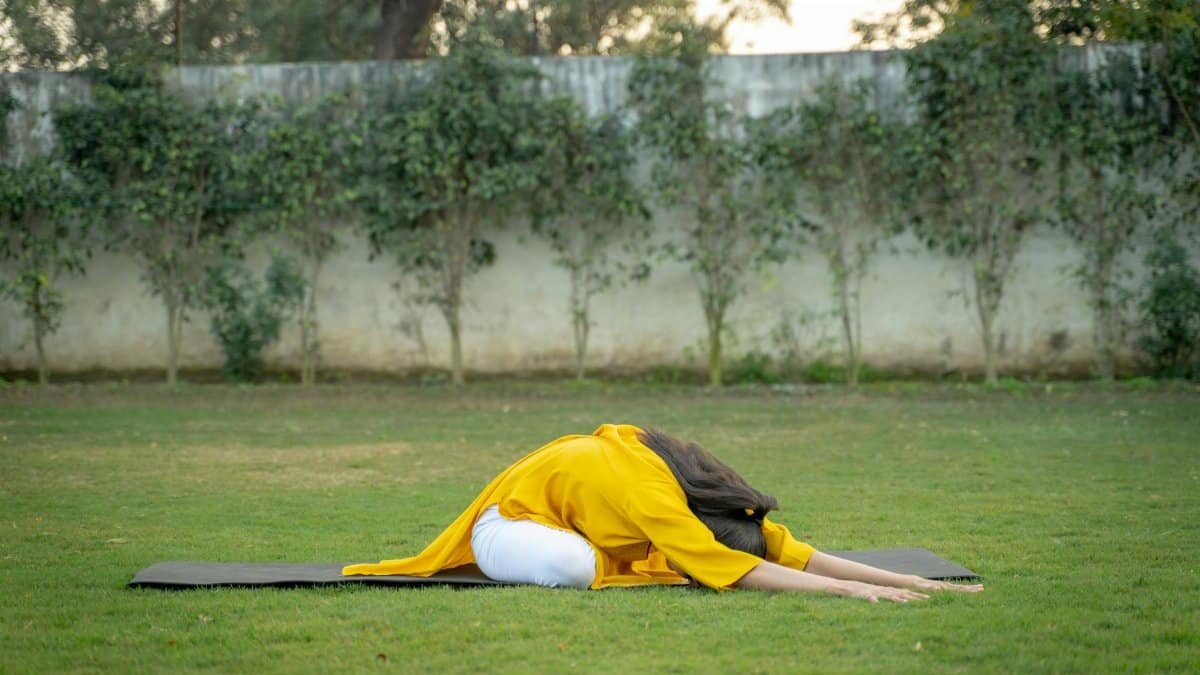 Adult woman performing yoga pose outdoors on a mat surrounded by green grass and trees.