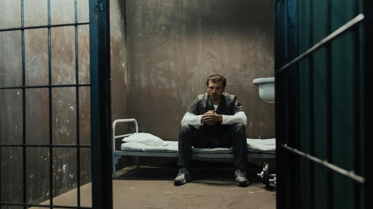 A man sits alone, reflecting inside a dimly lit prison cell.