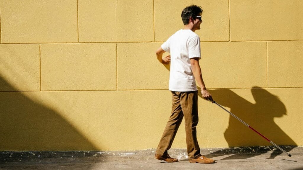 Side view of a blind man outdoors walking with a cane along a sunlit wall.