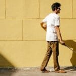 Side view of a blind man outdoors walking with a cane along a sunlit wall.