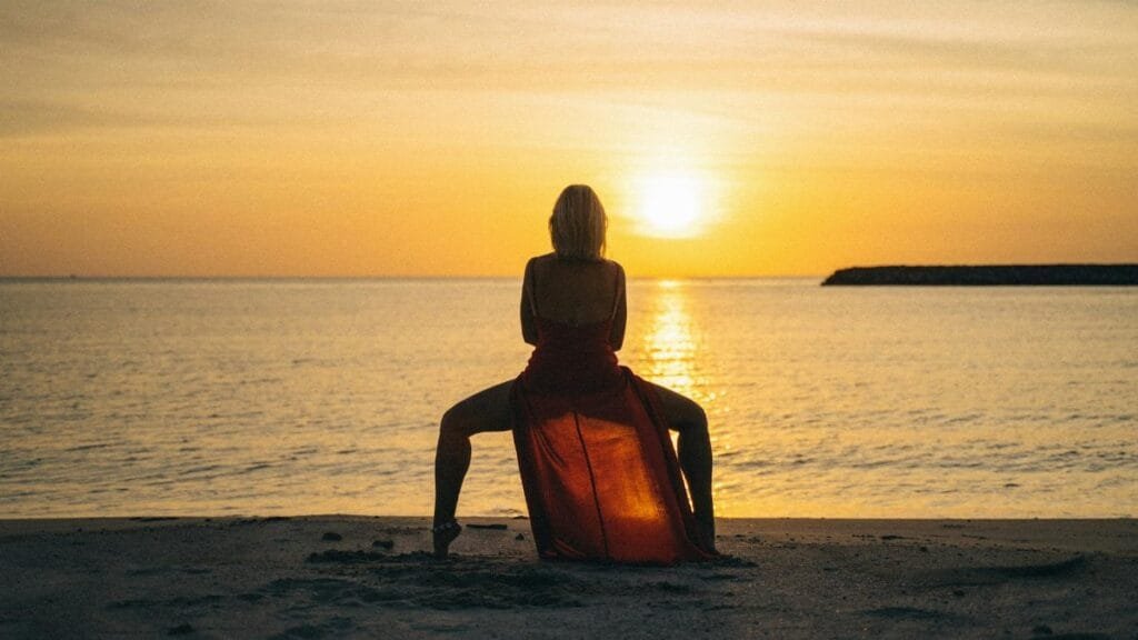 A woman in a red dress performs yoga at sunset on a serene beach.