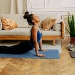 Woman performing yoga indoors on a mat with laptop nearby for an online session.