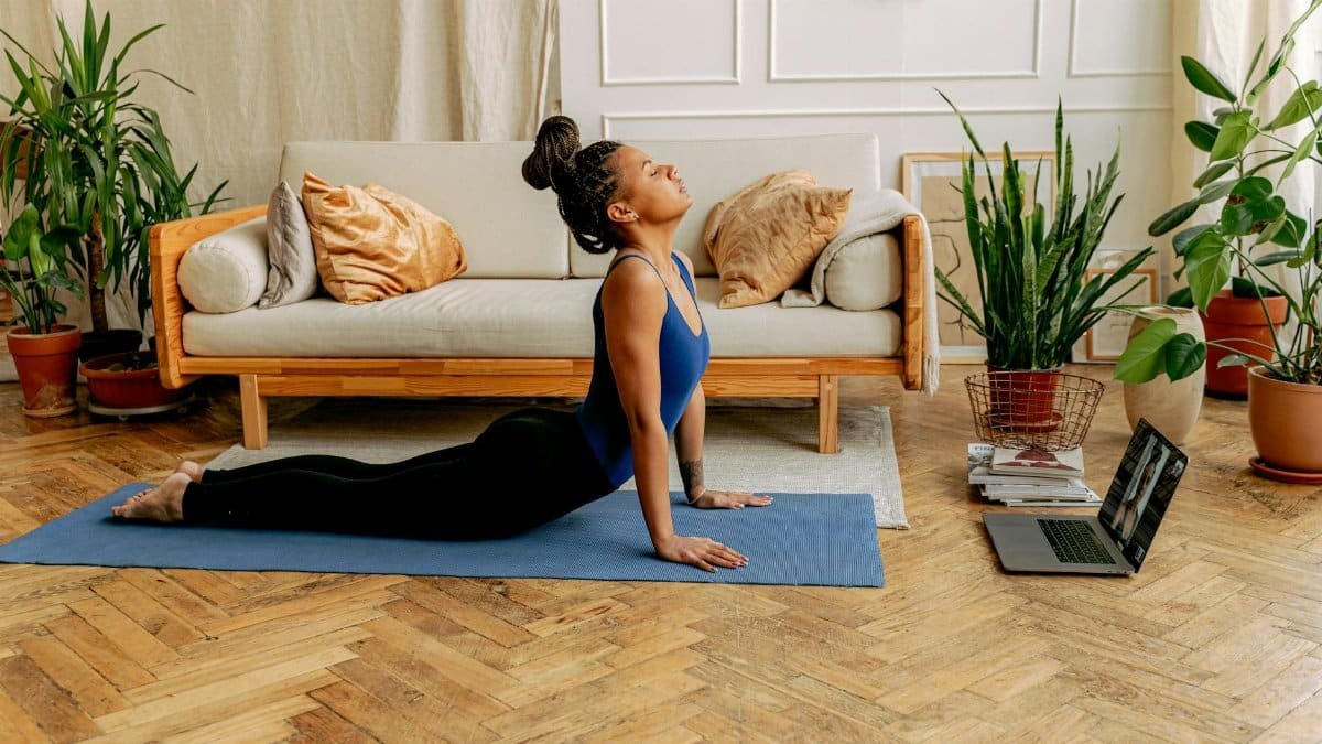 Woman performing yoga indoors on a mat with laptop nearby for an online session.