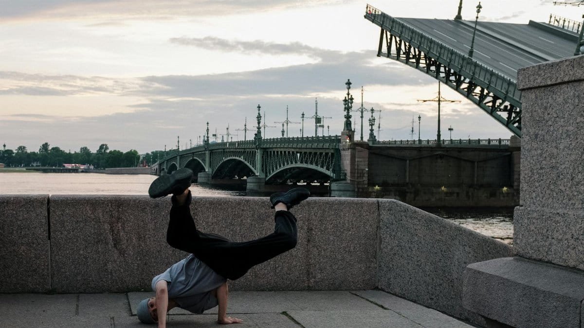 A man performs a dynamic breakdance move by a raised bridge at dusk in the city.