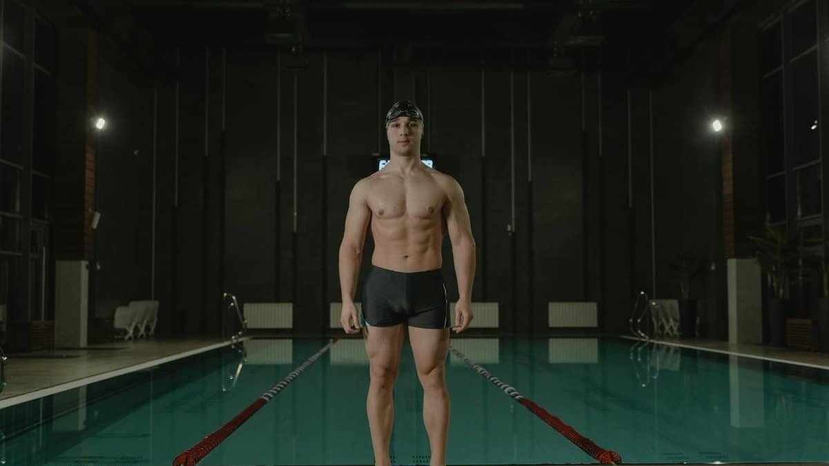 Athletic male swimmer standing confidently poolside indoors.