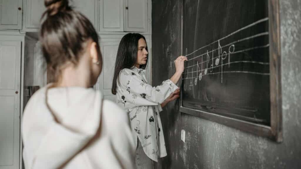 A student observes a teacher writing music notes on a classroom blackboard.