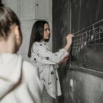 A student observes a teacher writing music notes on a classroom blackboard.