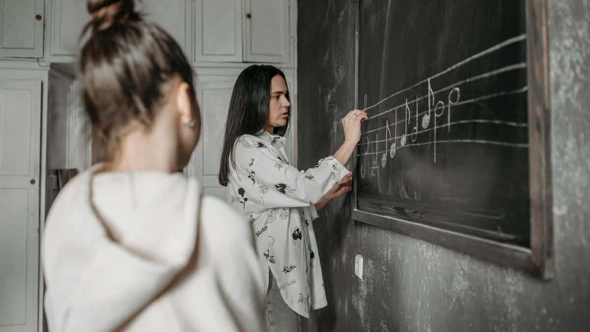 A student observes a teacher writing music notes on a classroom blackboard.