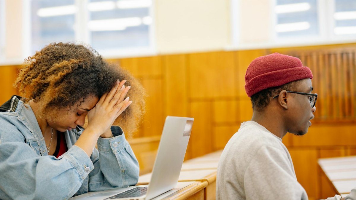 Two students working on laptops in a classroom, showing academic stress.
