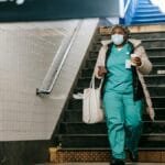 Full body calm adult African American nurse in outerwear and face mask walking with coffee to go in New York underground station