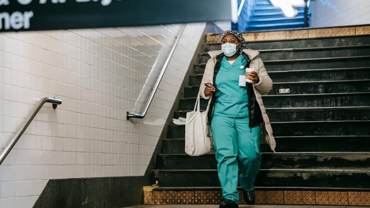 Full body calm adult African American nurse in outerwear and face mask walking with coffee to go in New York underground station