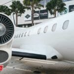 Private jet parked at a modern airport terminal with palm trees and shopping signs visible.