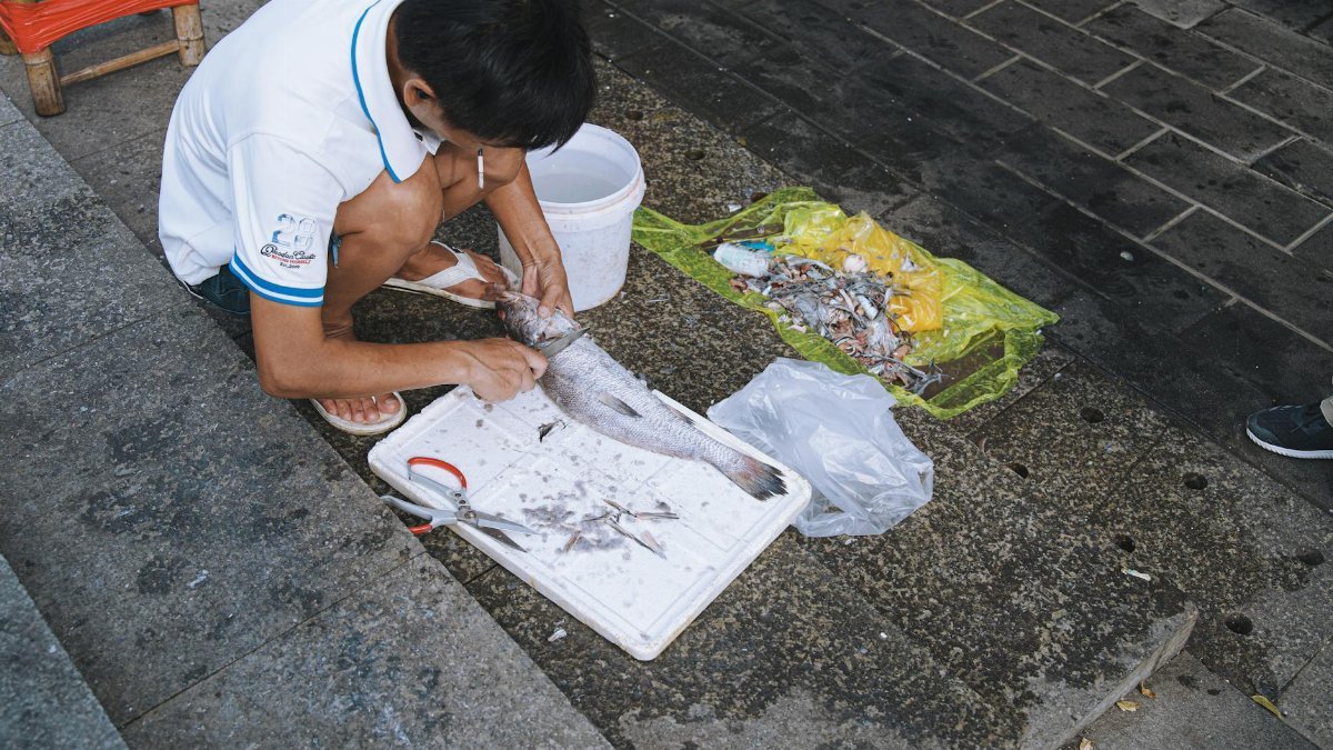A man crouches on steps scaling fish, surrounded by urban textures and waste.