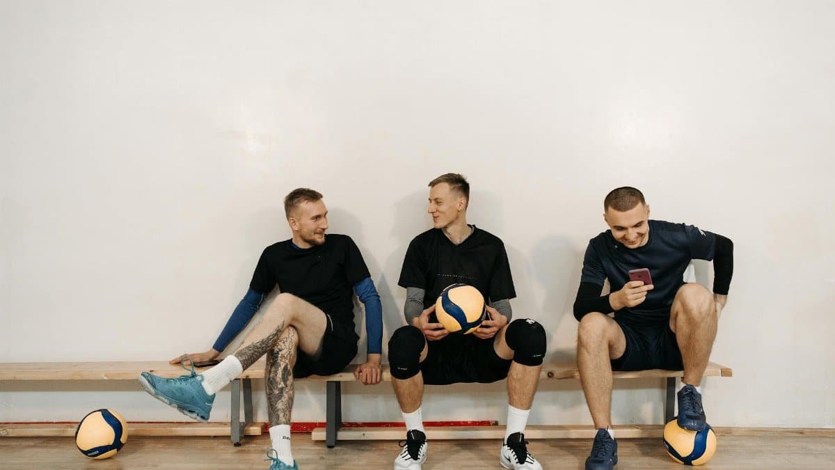 Three male athletes sitting on a bench with volleyballs, sharing a conversation indoors.