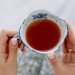 A detailed view of hands holding an ornate ceramic tea cup with warm tea indoors.