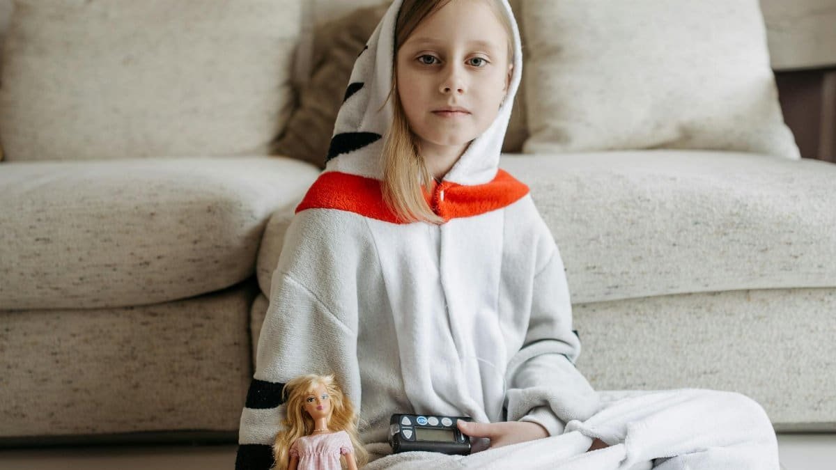 Young girl in onesie sitting at home with an insulin pump and doll, illustrating diabetes management.