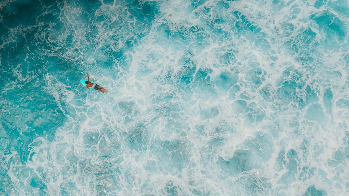Drone captures a surfer paddling through vibrant blue ocean waves, surrounded by swirling foam.
