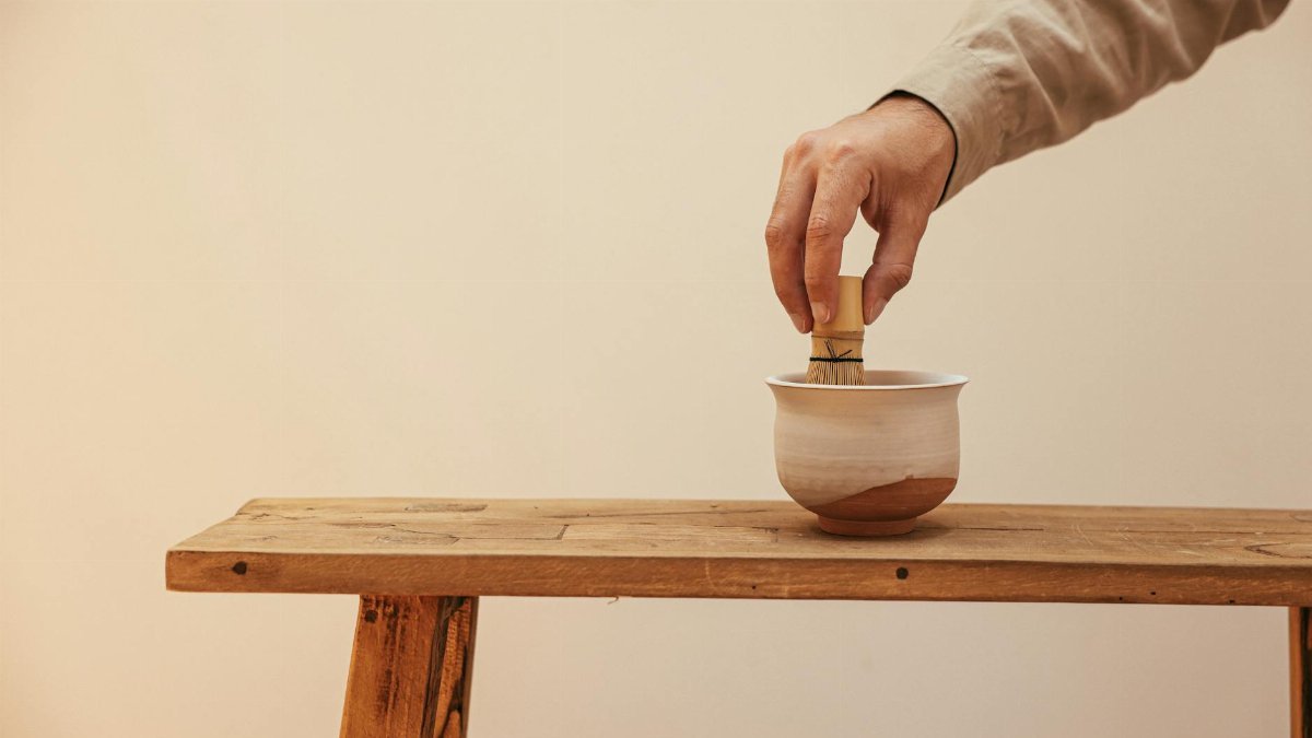 Calm tea ceremony scene with a hand whisking tea in a ceramic cup on a wooden bench.