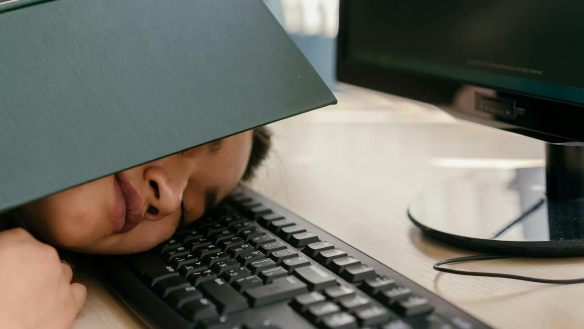 A young woman falls asleep on a keyboard with a textbook on her face, depicting study fatigue.