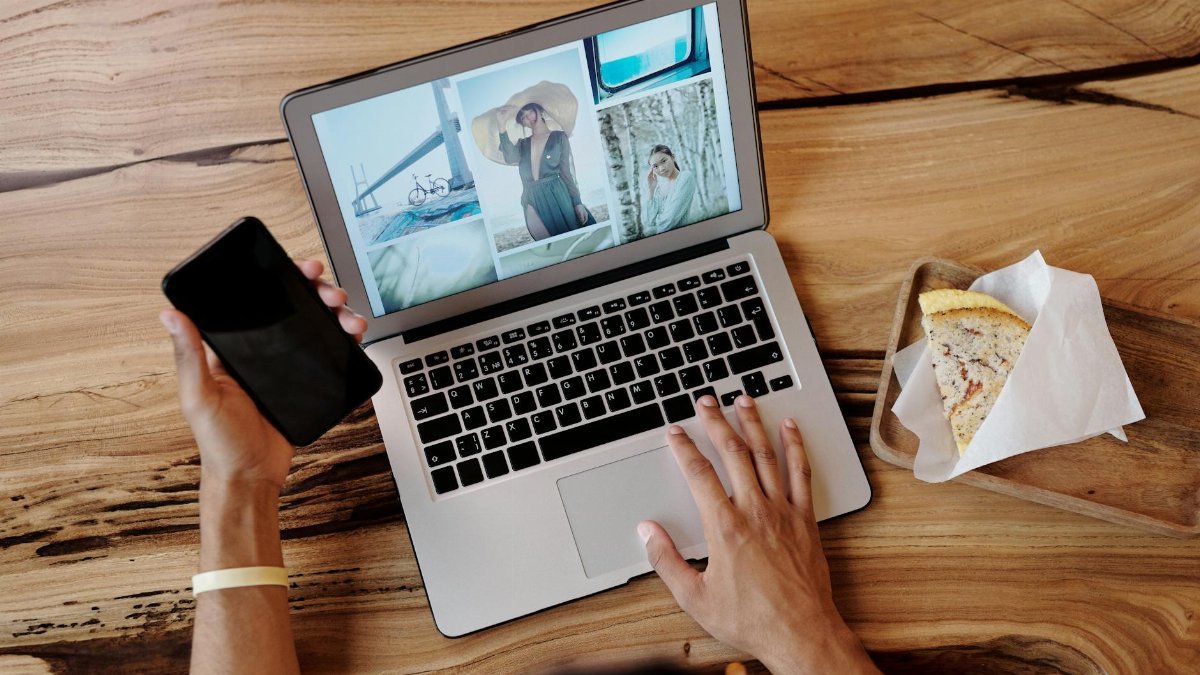 Person uses smartphone and laptop while enjoying a sandwich at a wooden table, showcasing multitasking and technology use.