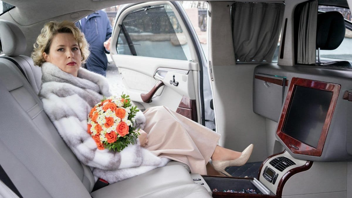 Cheerful female with curly hair wearing dress and fur coat sitting in passenger seat of luxurious car with opened door with bouquet of colorful flowers