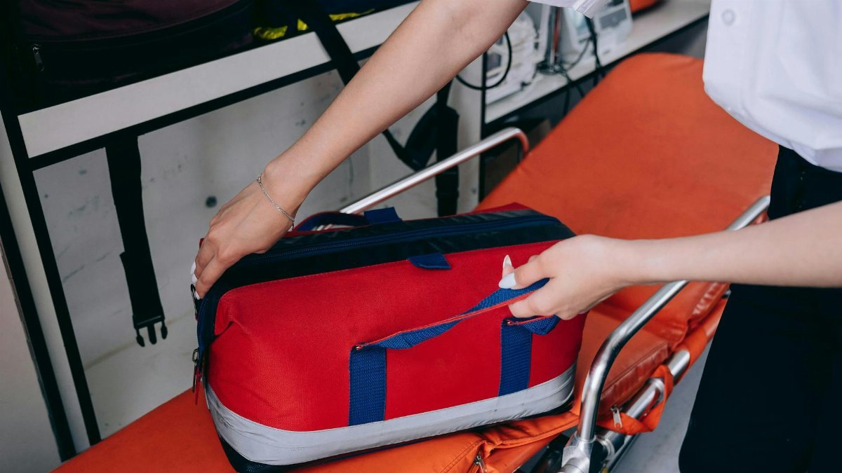A healthcare worker reaches for a red medical bag on a stretcher indoors.