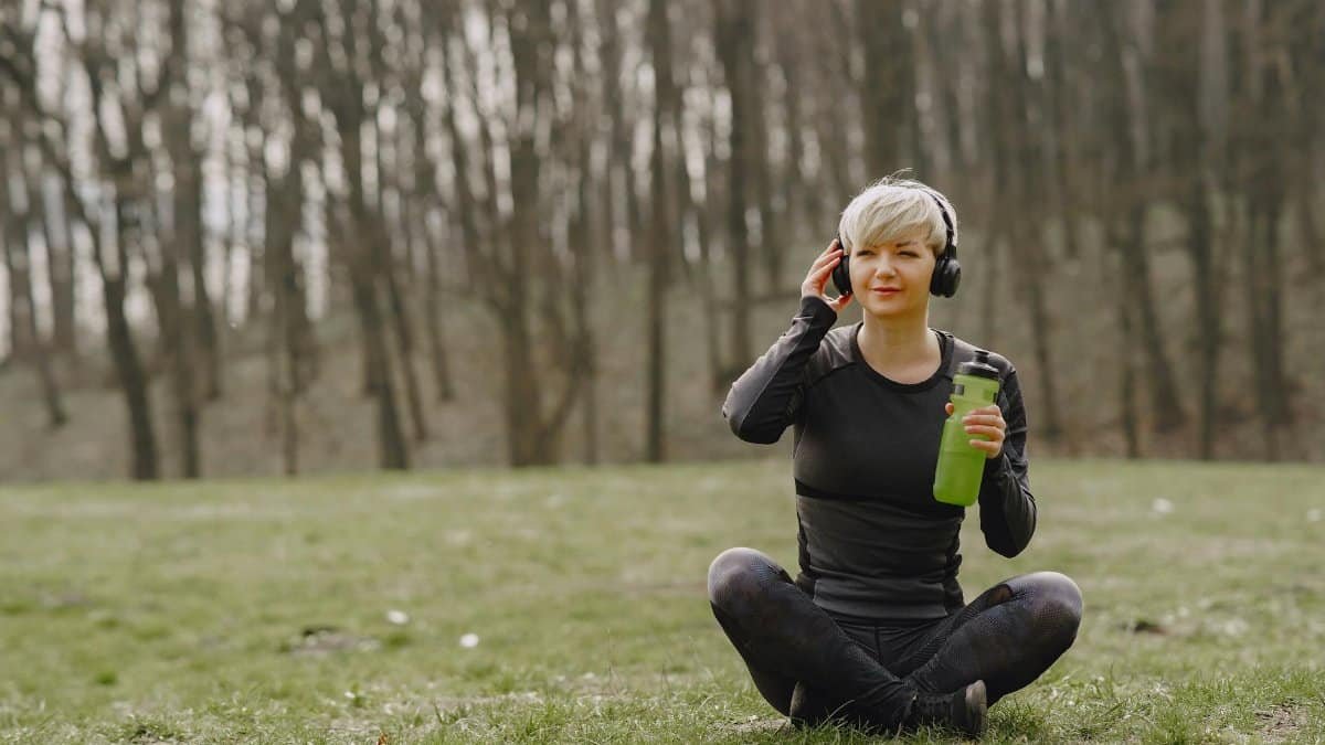 Woman sitting cross-legged in forest park with headphones and water bottle, enjoying a peaceful moment.