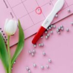 A close-up of a pregnancy test, calendar, and tulip arranged on a pink surface.